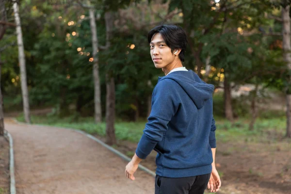 Image of smiling asian guy in casual wear and eyeglasses walking down road through park with green trees