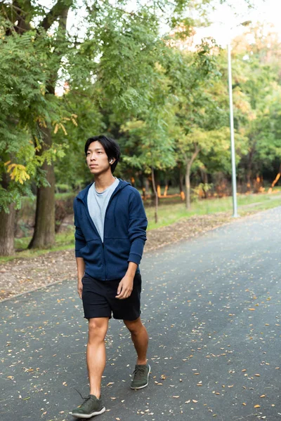 Image of pleased asian guy in casual wear and eyeglasses walking down road through park with green trees