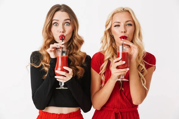两个开朗的年轻穿着漂亮的妇女化妆站在白色背景隔离 喝番茄汁从眼镜的肖像的肖像 — 图库照片