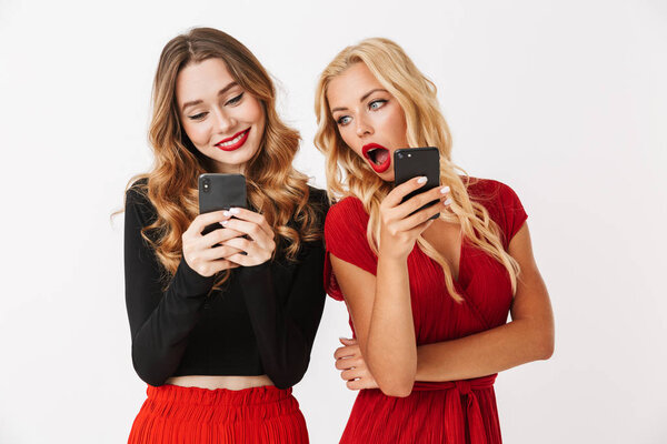 Portrait of two excited young smartly dressed women wearing makeup standing isolated over white background, looking at each other mobile phones