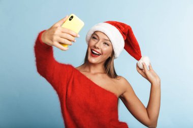 Mavi duvar arka plan üzerinde izole poz güzel duygusal heyecanlı genç kadın giyen Noel şapka görüntüsünü almak bir selfie cep telefonuyla.