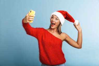 Mavi duvar arka plan üzerinde izole poz güzel duygusal heyecanlı genç kadın giyen Noel şapka görüntüsünü almak bir selfie cep telefonuyla.
