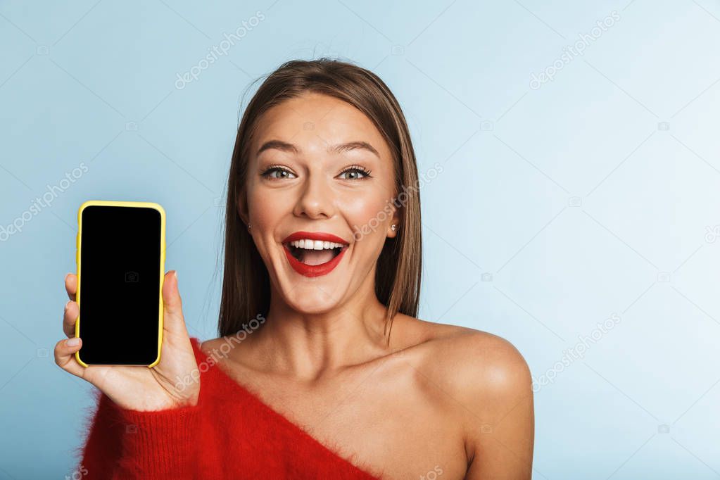 Image of a beautiful excited young woman posing isolated over blue wall background using mobile phone showing empty display.