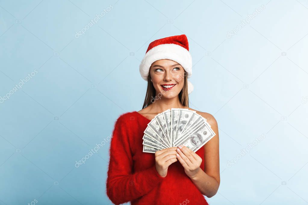 Cheerful young woman wearing santa claus hat standing isolated over blue background, showing money banknotes