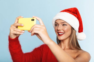Mavi duvar arka plan üzerinde izole poz güzel duygusal heyecanlı genç kadın giyen Noel şapka görüntüsünü almak bir selfie cep telefonuyla.