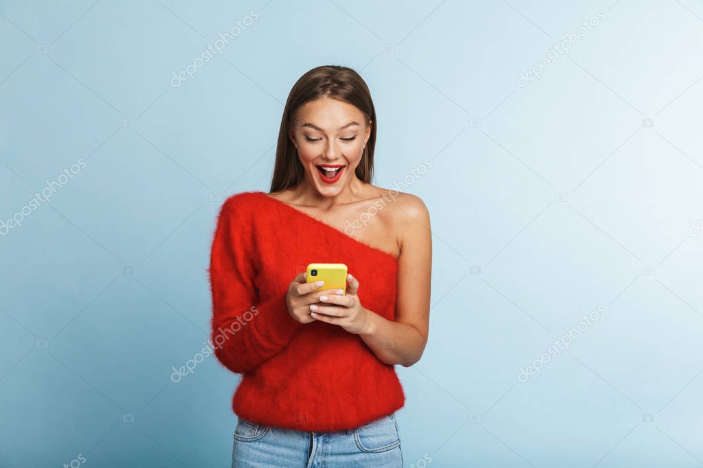 Image of a beautiful emotional excited young woman posing isolated over blue wall background using mobile phone.