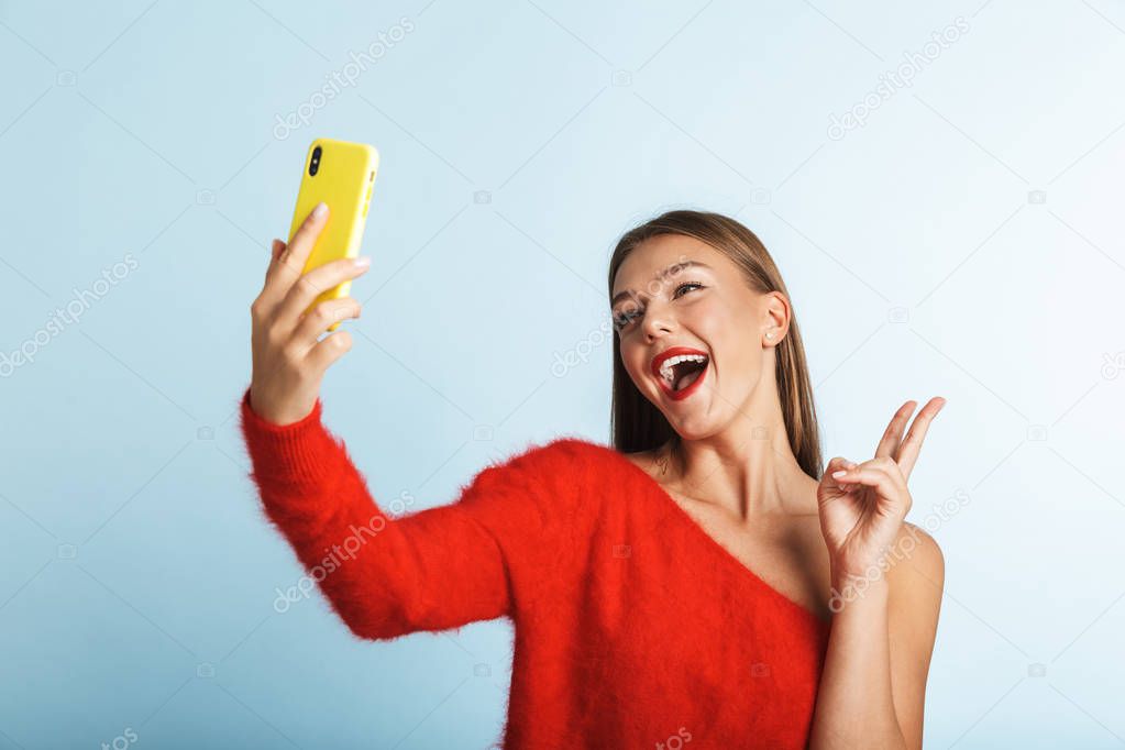 Image of a beautiful emotional excited young woman posing isolated over blue wall background take a selfie by mobile phone.