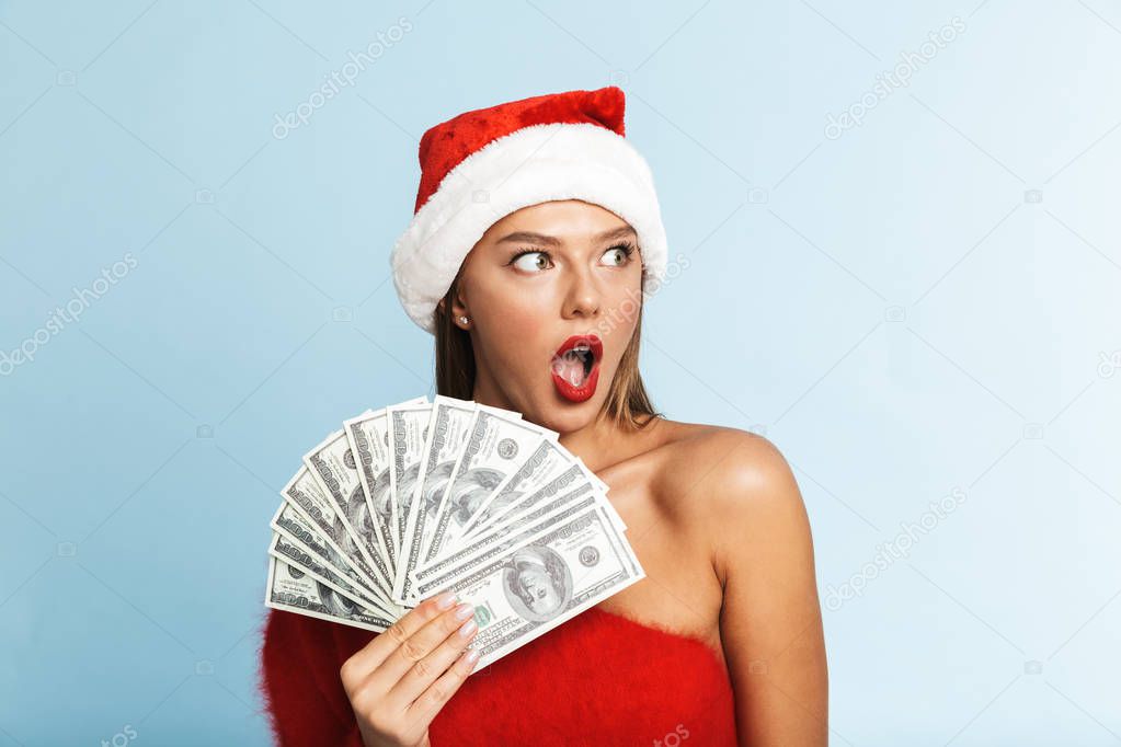 Image of a beautiful happy young woman wearing christmas hat posing isolated over blue wall background holding money.