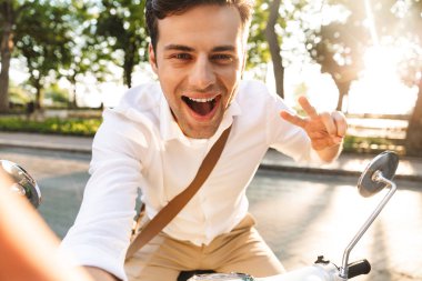 Açık havada scooter üzerinde yürüyen bir yakışıklı heyecanlı genç iş adamı fotoğrafını çekin bir selfie barış gösterilen kamera tarafından.
