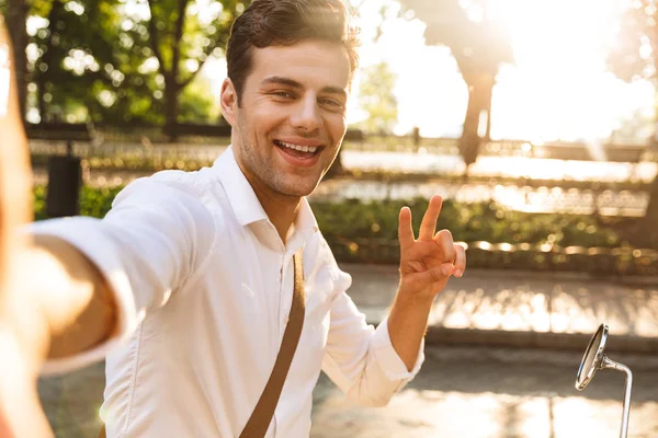 スクーターで野外を歩いているハンサムな興奮若いビジネス人の写真を取る Selfie 平和を示すカメラ — ストック写真