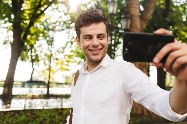 Outsretched の手で携帯電話を保持している都市公園で屋外に立っている間 Selfie を取ってシャツを着て幸せな青年実業家 — ストック写真