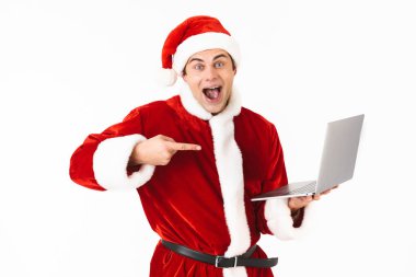 Neşeli bir genç adam portresi beyaz arka plan üzerinde izole ayakta, dizüstü bilgisayar tutan, parmakla Noel Baba kostümü giymiş