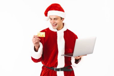 Heyecanlı bir genç adam portresi dizüstü bilgisayar, plastik kredi kartı gösteren holding beyaz arka plan üzerinde izole ayakta Noel Baba kostüm giymiş
