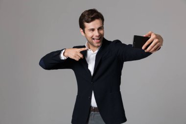 Görüntü gri arka plan üzerinde izole Smartphone cep telefonu ile selfie fotoğraf çekerken gülüyor takım elbiseli yakışıklı işadamı 30'lu