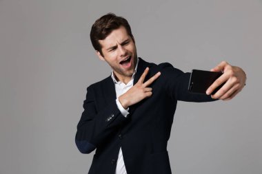 Mutlu işadamı 30'lu resmi kıyafetli barış işareti gri arka plan üzerinde izole cep telefonuyla selfie fotoğraf çekerken gösterilen görüntü