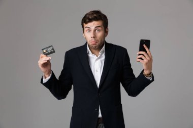 Görüntü kredi kartı ve cep telefonu gri arka plan üzerinde izole tutan takım elbiseli şaşkın işadamı 30'lu