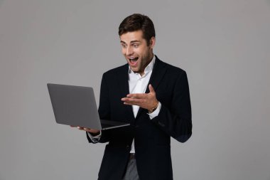 Dizüstü bilgisayar kullanan gri arka plan üzerinde izole takım elbise giymiş bir heyecanlı genç işadamı portresi