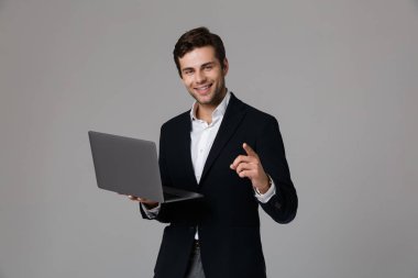 Görüntü gri arka plan üzerinde izole laptop kullanırken sevindirici takım elbiseli yakışıklı işadamı 30'lu