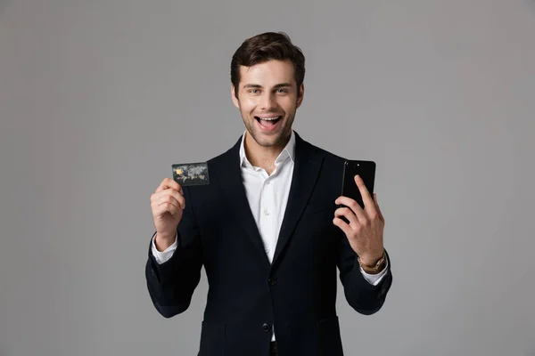 穿着西装的30岁男子的形象 手持黑色手机和信用卡 在灰色背景中隔离 — 图库照片