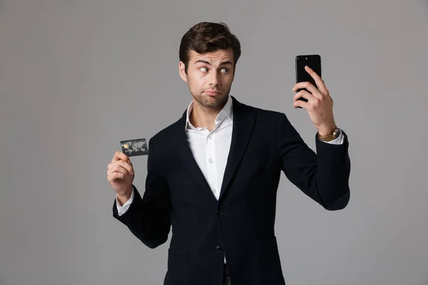 穿着西装的30多岁帅哥的形象 拿着黑色手机和信用卡 在灰色背景中被隔离 — 图库照片