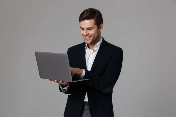 灰色の背景の上分離されたノート パソコンを使用しながら歓喜のスーツで満足している実業家 代のイメージ — ストック写真