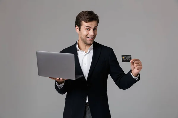 明るいビジネスマンのイメージ30代スーツの笑顔を保持しながらノートパソコンとクレジットカードグレーの背景に隔離 — ストック写真