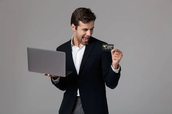 在灰色背景下拿着笔记本电脑和信用卡时 穿着西装微笑的快乐商人30多岁的形象 — 图库照片