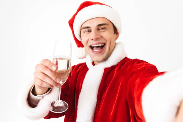英俊的男人30多岁的肖像在圣诞老人服装和红帽子拍照自拍照片 同时举行玻璃与香槟隔离在白色背景在工作室 图库图片