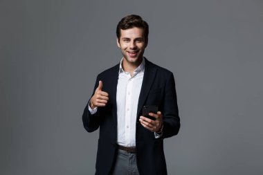 Mutlu bir genç işadamı portresi cep telefonu, jest Yaşasın gösterilen tutan gri arka plan üzerinde izole takım elbise giymiş