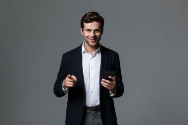 Mutlu bir genç işadamı portresi cep telefonu tutan, parmakla kameraya gri arka plan üzerinde izole takım elbise giymiş