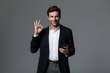 Neşeli bir genç işadamı portresi cep telefonu, ok jest gösterilen holding, gri arka plan üzerinde izole takım elbise giymiş