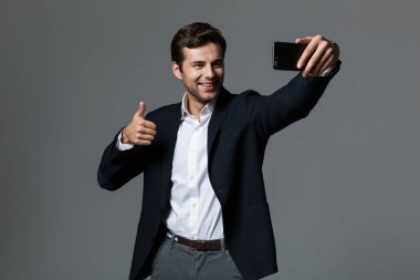 Neşeli bir genç işadamı portresi bir selfie alarak, gri arka plan üzerinde izole takım elbise giymiş