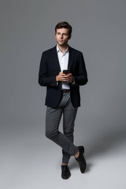 Yakışıklı bir genç işadamı portresi tam uzunlukta cep telefonu holding ayakta, gri arka plan üzerinde izole takım elbise giymiş