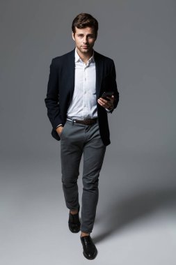 Kendine güvenen genç işadamı portresi tam uzunlukta cep telefonu tutan yürüme, gri arka plan üzerinde izole takım elbise giymiş