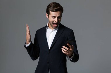 Mutlu bir genç işadamı portresi giyinmiş elbiseli cep telefonu, holding, gri arka plan üzerinde izole kutlama