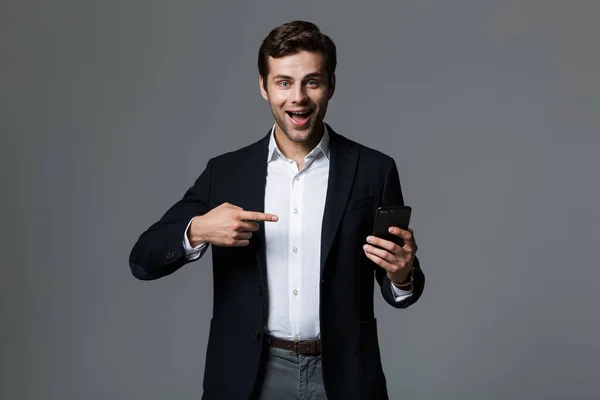 一个开朗的年轻商人的肖像 穿着西装 孤立在灰色的背景下 指着手机指指点点 — 图库照片