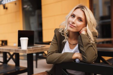 Paket servisi olan restoran kahve ve gümüş laptop ile açık kafede otururken kameraya gülümseyen genç sarışın bir kadın portresi