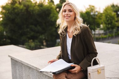 Görüntü gülümseyen ve beton parapet açık otururken kağıt klasör tutan büyüleyici kadın
