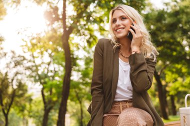 Sarışın kadın 20'li yaşlarda görüntüsünü parkta bankta otururken çağrısı için smartphone kullanarak