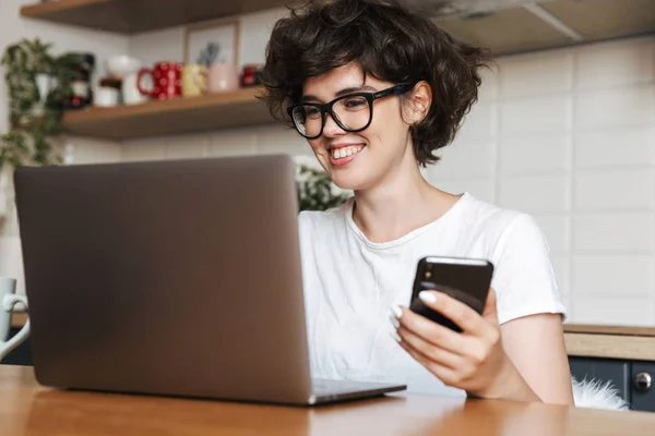 一个快乐的年轻女子的肖像戴眼镜在家里的笔记本电脑工作 拿着手机 — 图库照片