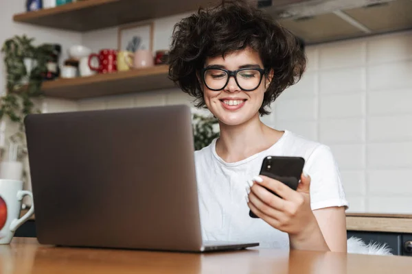 一个快乐的年轻女子的肖像戴眼镜在家里的笔记本电脑工作 拿着手机 — 图库照片