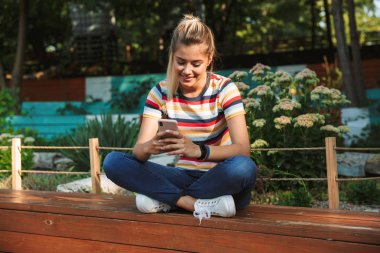 Parkta bir bankta oturmuş, seyir tmobile telefon, gülen bir mutlu genç Genç kız portresi