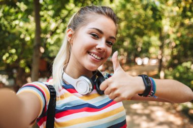 Mutlu sevimli genç kız görüntüsünü almak bir selfie kamera Park açık havada başparmak gösterilmesini.