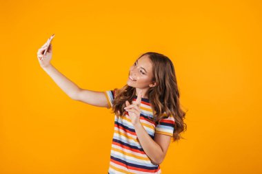 Sarı duvar arka plan üzerinde poz mutlu bir genç kızın görüntü almak bir selfie cep telefonuyla.