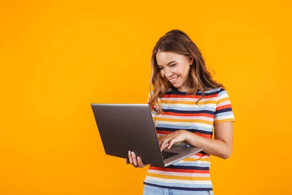 一个微笑的开朗的年轻女孩的肖像站在孤立的黄色背景 使用笔记本电脑 — 图库照片