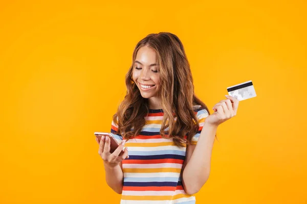 笑みを浮かべて陽気な若い少女が立っている孤立した黄色の背景の上の携帯電話を使用してプラスチック製のクレジット カードを示す肖像画 — ストック写真