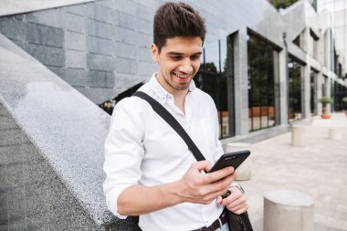Gülümseyen başarılı genç işadamı açık havada yürüyüş, cep telefonuyla beyaz gömlek giymiş