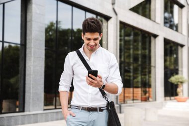 Gülümseyen başarılı genç işadamı açık havada yürüyüş, cep telefonuyla beyaz gömlek giymiş