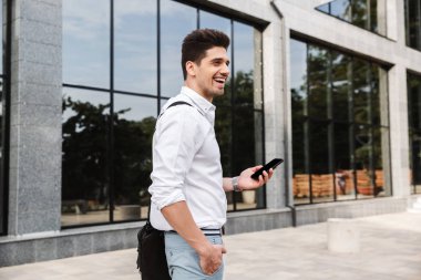 Gülümseyen başarılı genç işadamı açık havada yürüyüş, cep telefonuyla beyaz gömlek giymiş