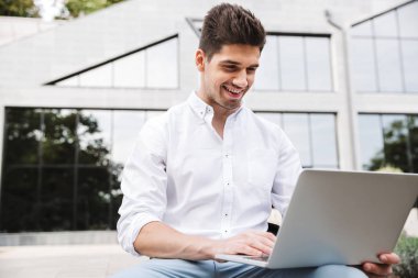 Açık havada otururken dizüstü bilgisayarda çalışarak gülümseyen genç iş adamı